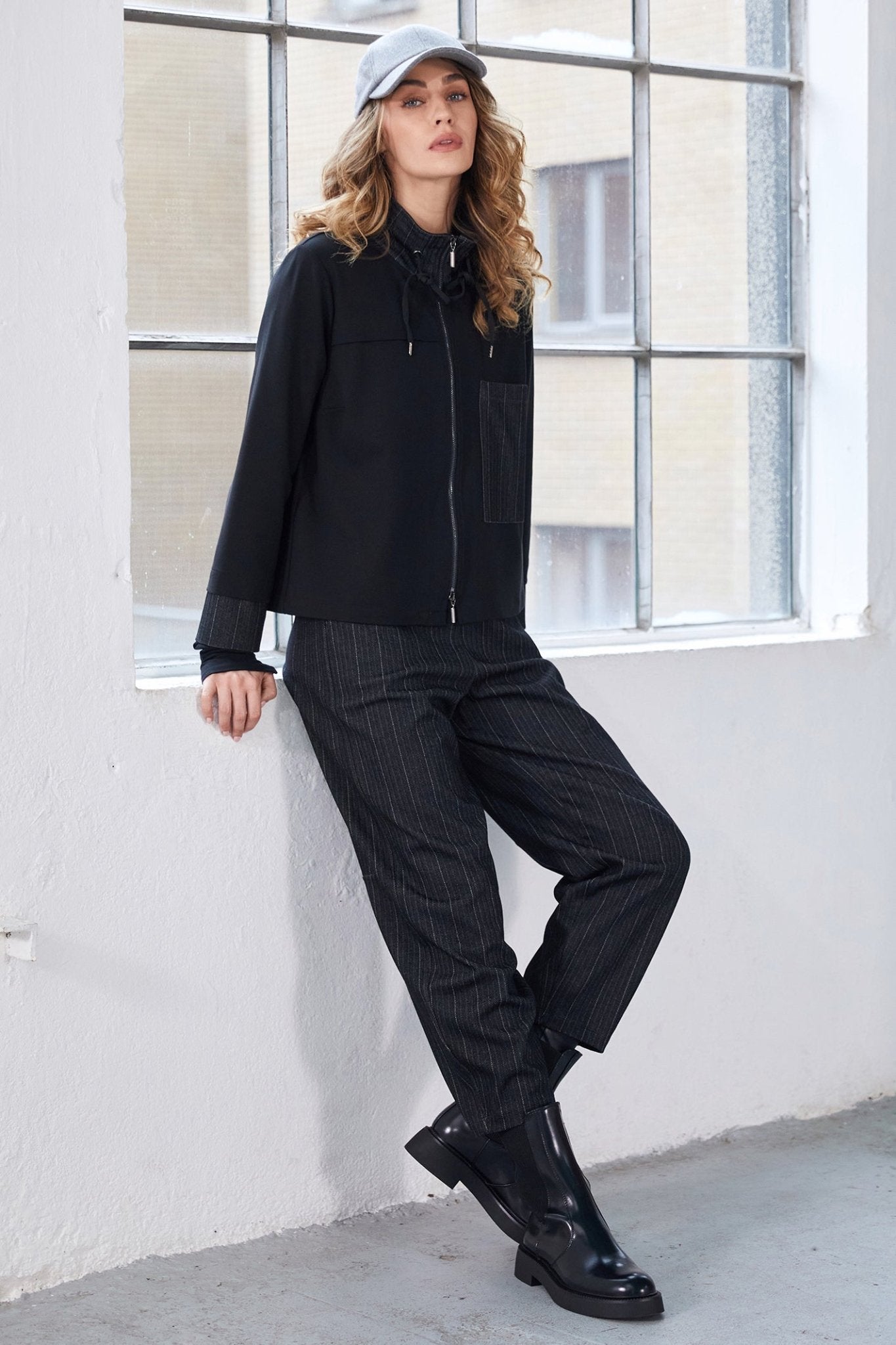 Person wearing a black jacket, pants, and shoes leaning against a white wall with large windows.