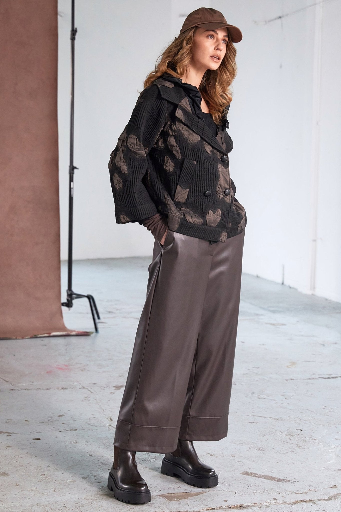 Woman wearing a black patterned coat, brown pants, and a cap in a studio setting.