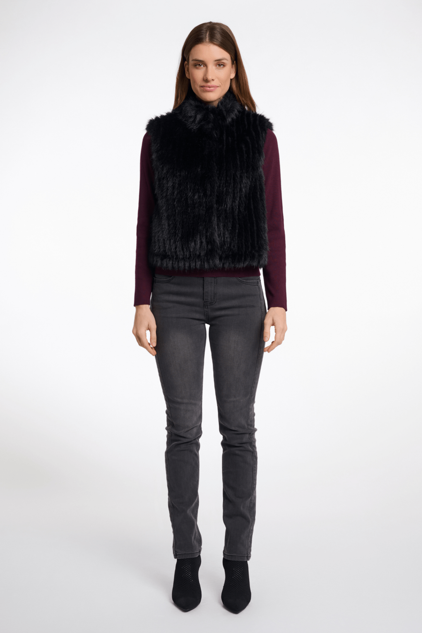 Woman wearing a black fur vest, purple top, gray jeans, and black boots on a white background