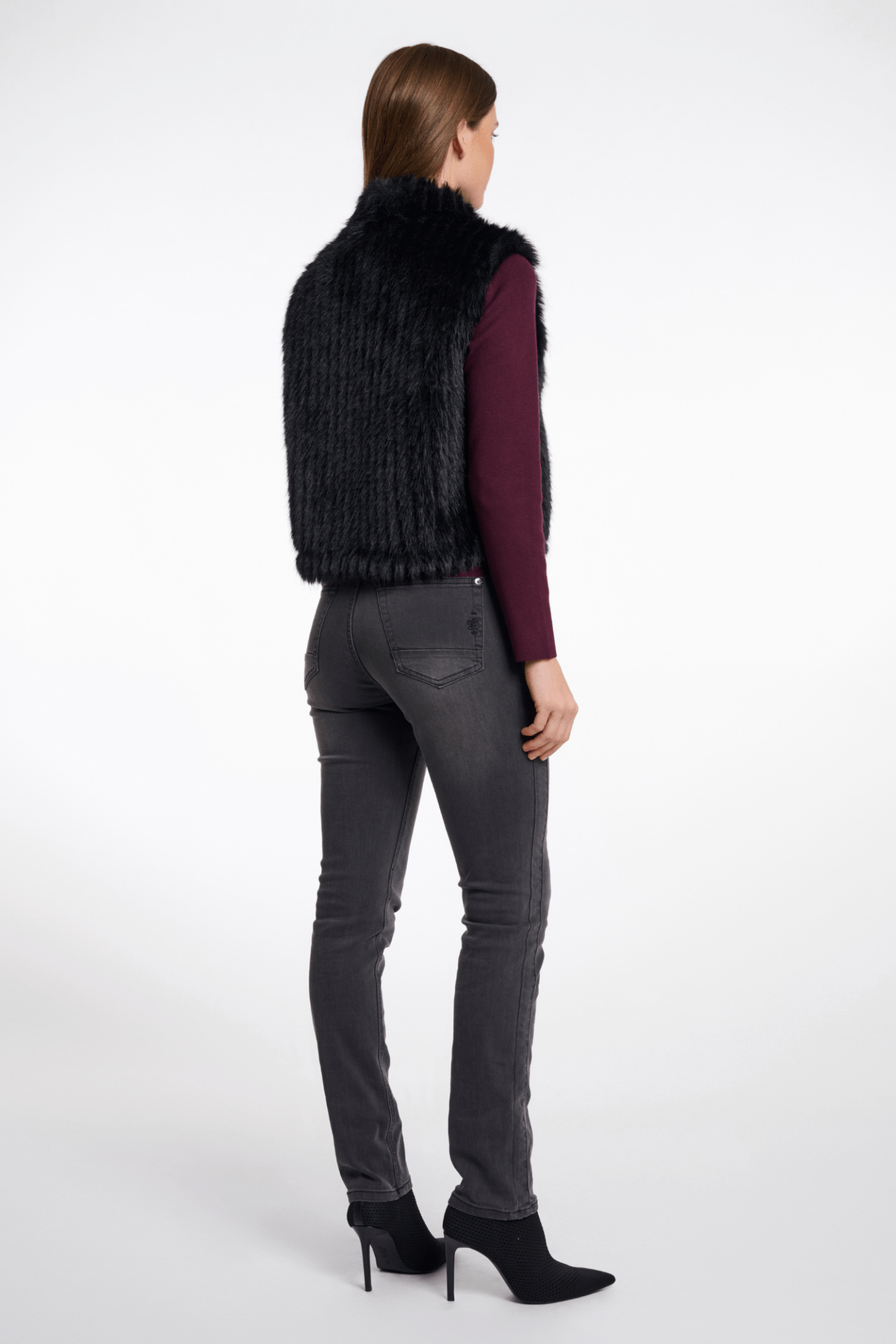 Person wearing a black fur vest, purple shirt, gray jeans, and black high-heeled shoes on a white background