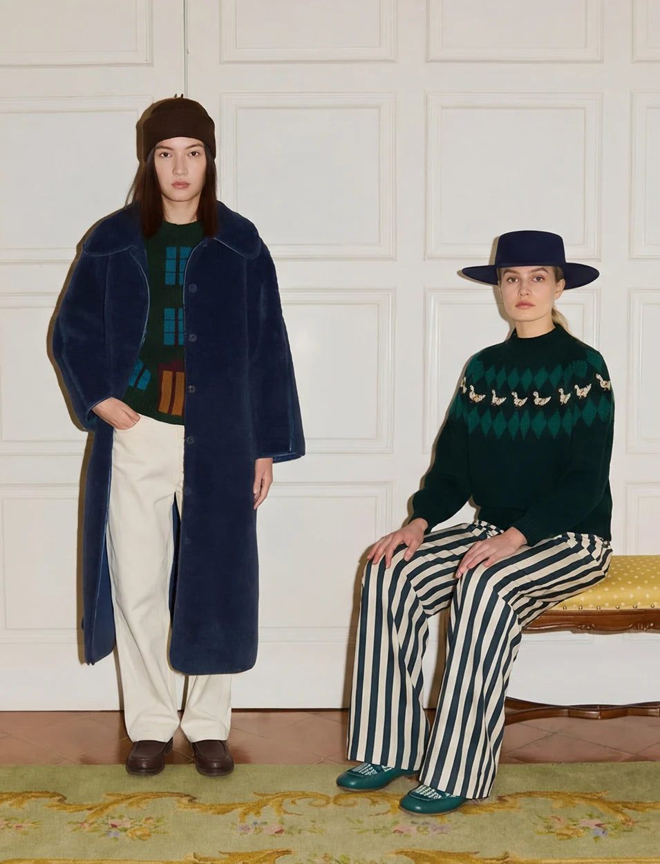 Two models posing in a styled indoor setting. The standing model wears a long navy blue coat over a green and blue patterned sweater, cream trousers, and a dark brown beret.