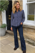 A woman wearing a navy and white vertically striped long-sleeve button-up shirt with dark blue jeans and brown shoes, standing on a stone patio in front of a house.