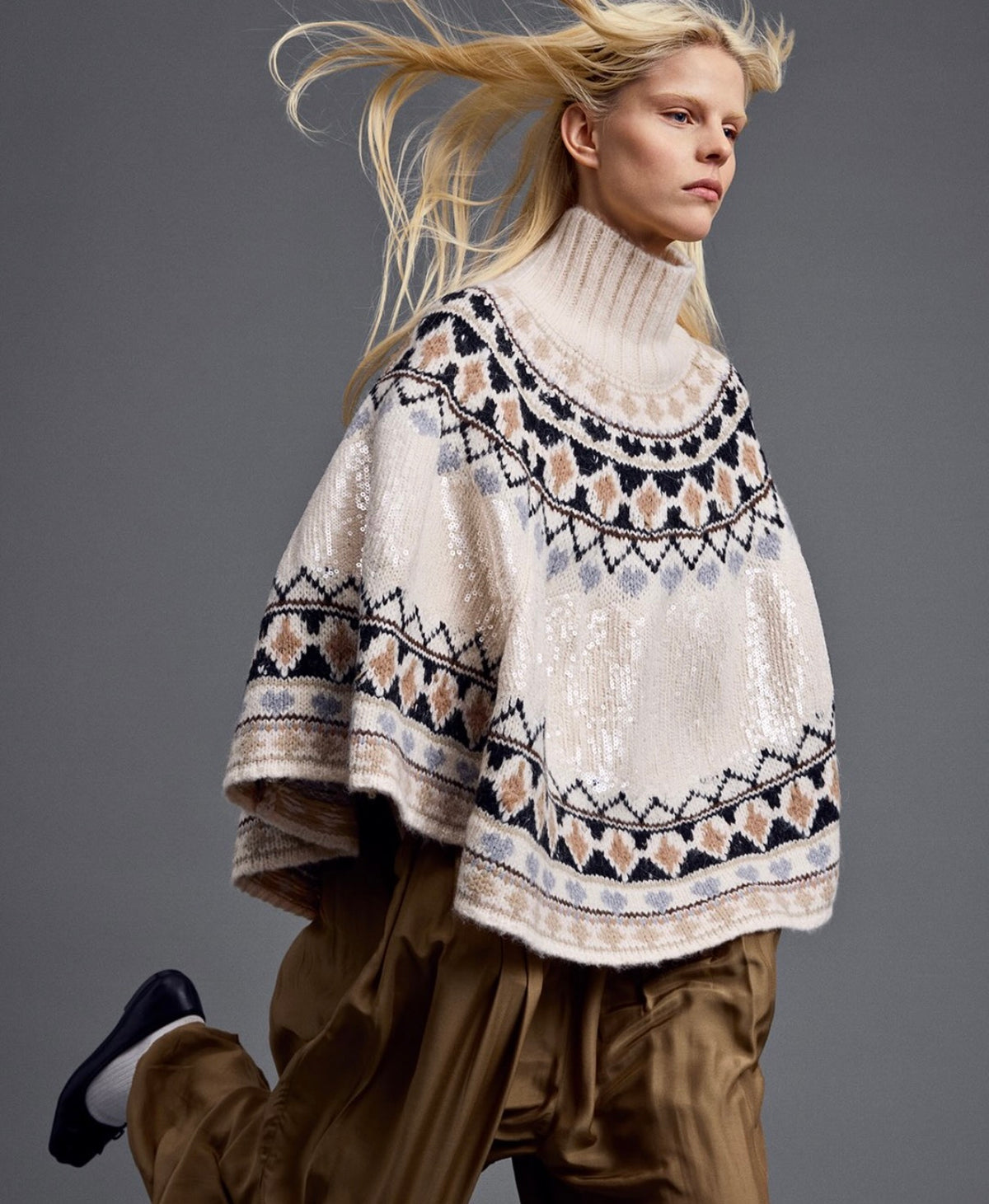 Woman wearing a cream patterned poncho with brown trousers against a gray background