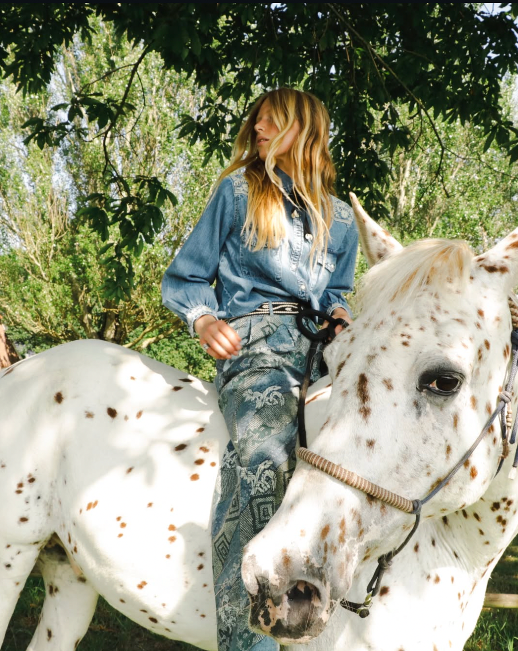 Woman riding a white spotted horse in a natural setting with trees.