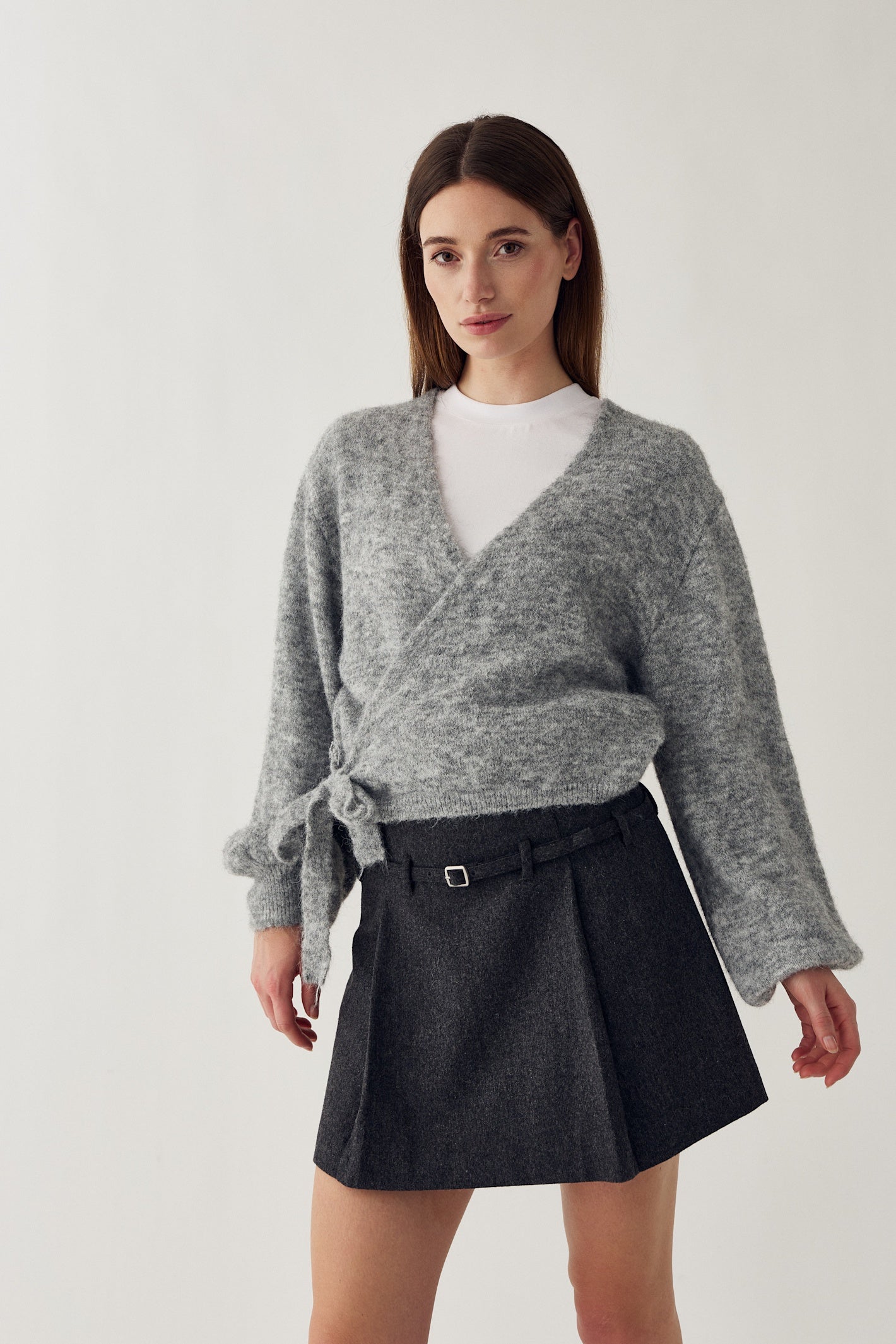 A woman modeling a heather gray wrap cardigan with a tie detail at the waist, worn over a white top and paired with a dark gray pleated mini skirt with a belt. Shop Noella at Dotique.