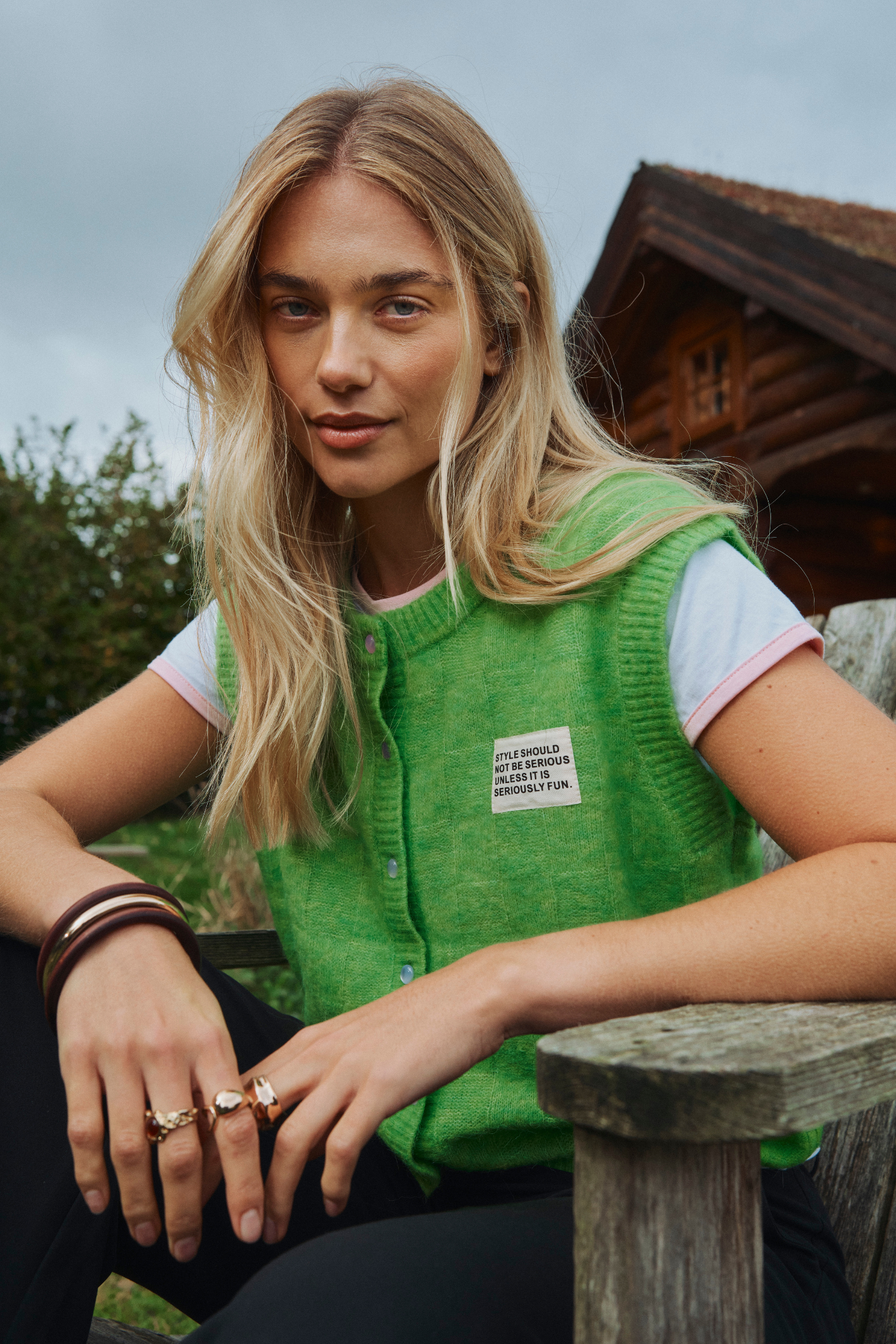 A woman wearing a bright green knit vest over a white t-shirt, sitting outdoors on a wooden bench with a rustic cabin in the background.