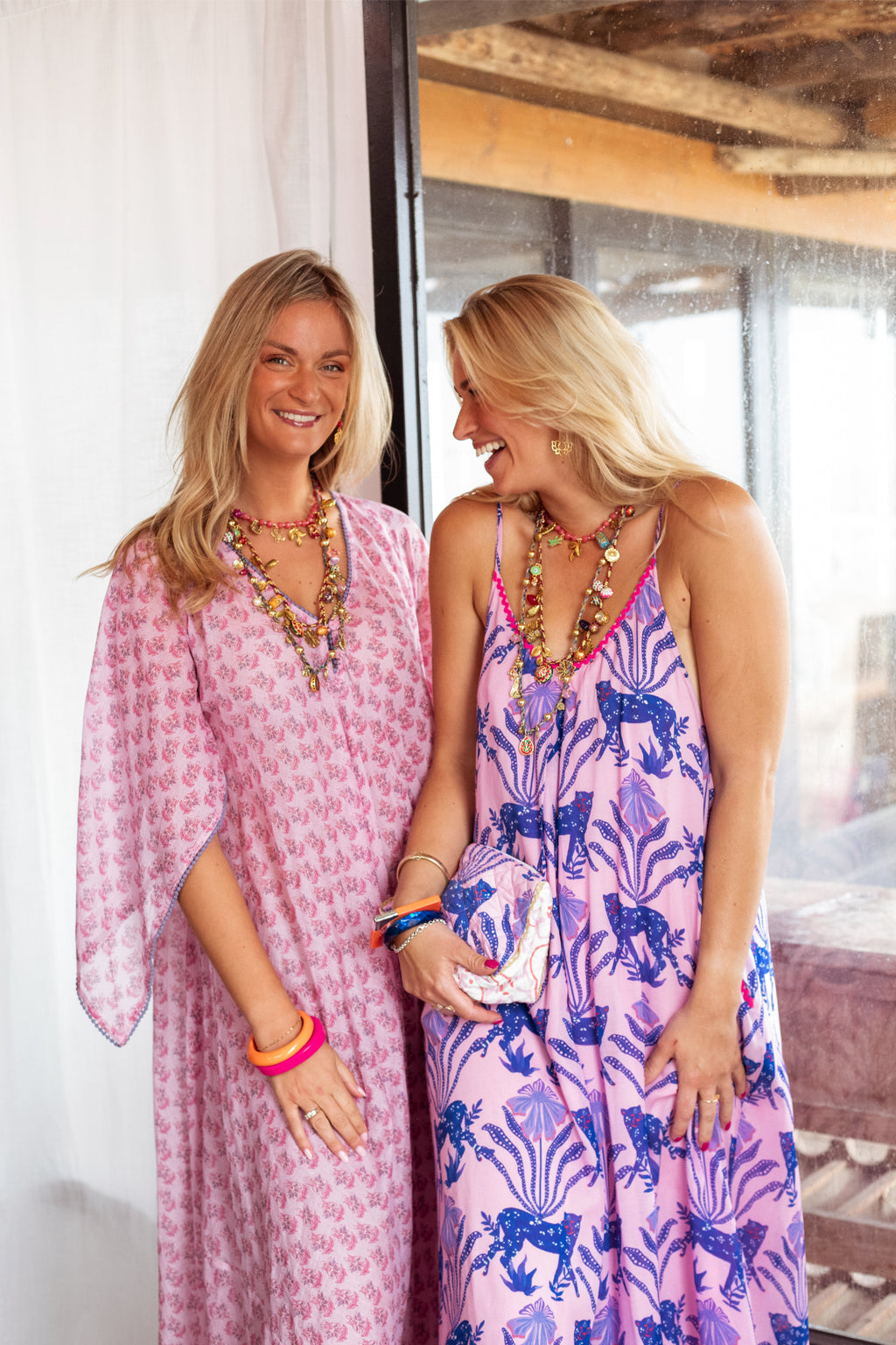 Two women standing next to each other, one in a pink dress and the other in a blue and white patterned dress, indoors.