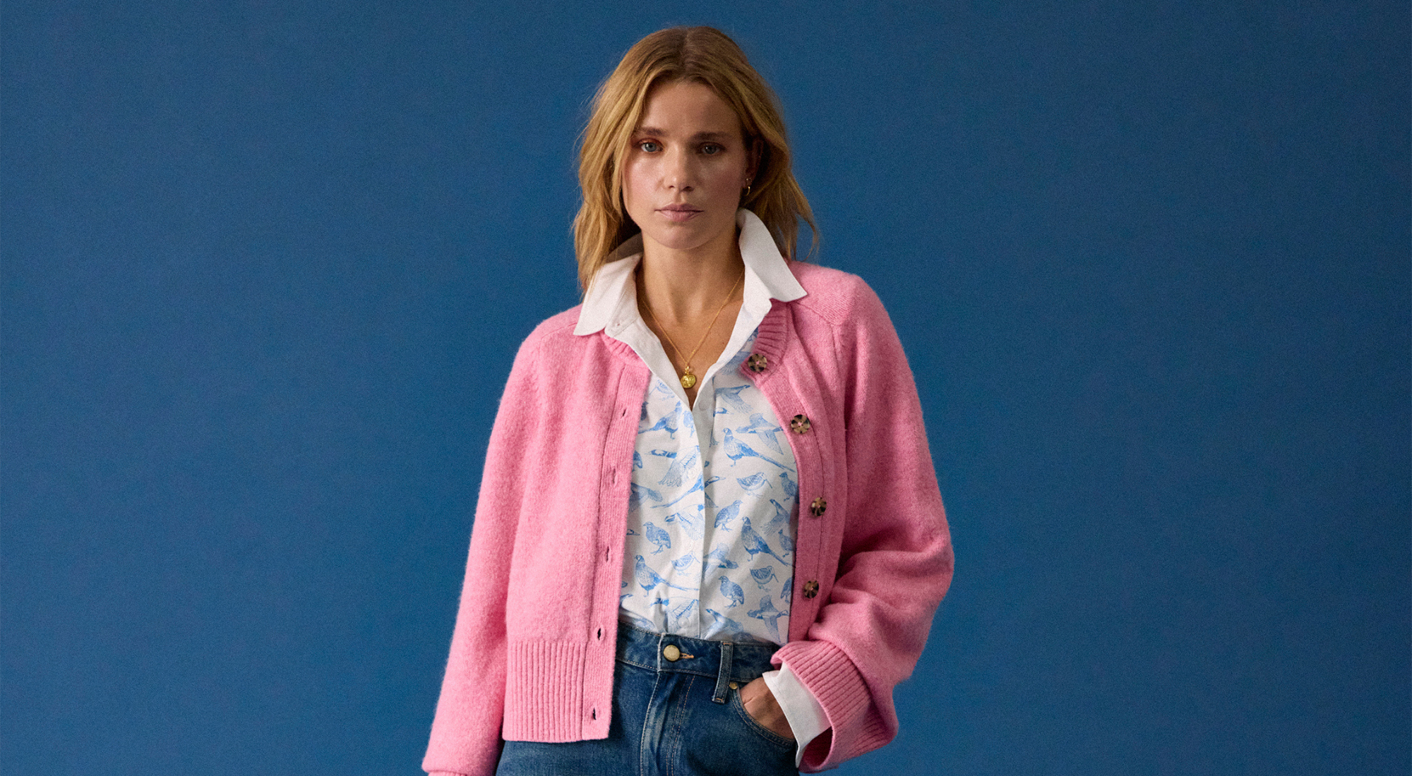 Woman wearing pink cardigan over white patterned blouse and blue jeans against solid blue backdrop Shop Dotique at Dotique