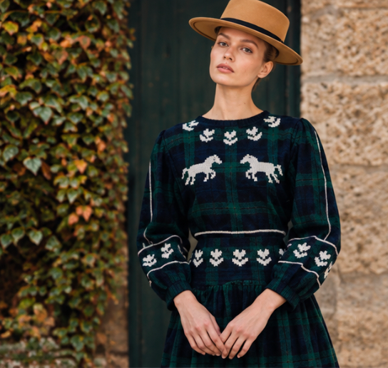Woman wearing a dark green tartan dress with white horse motifs and a tan fedora outdoors
