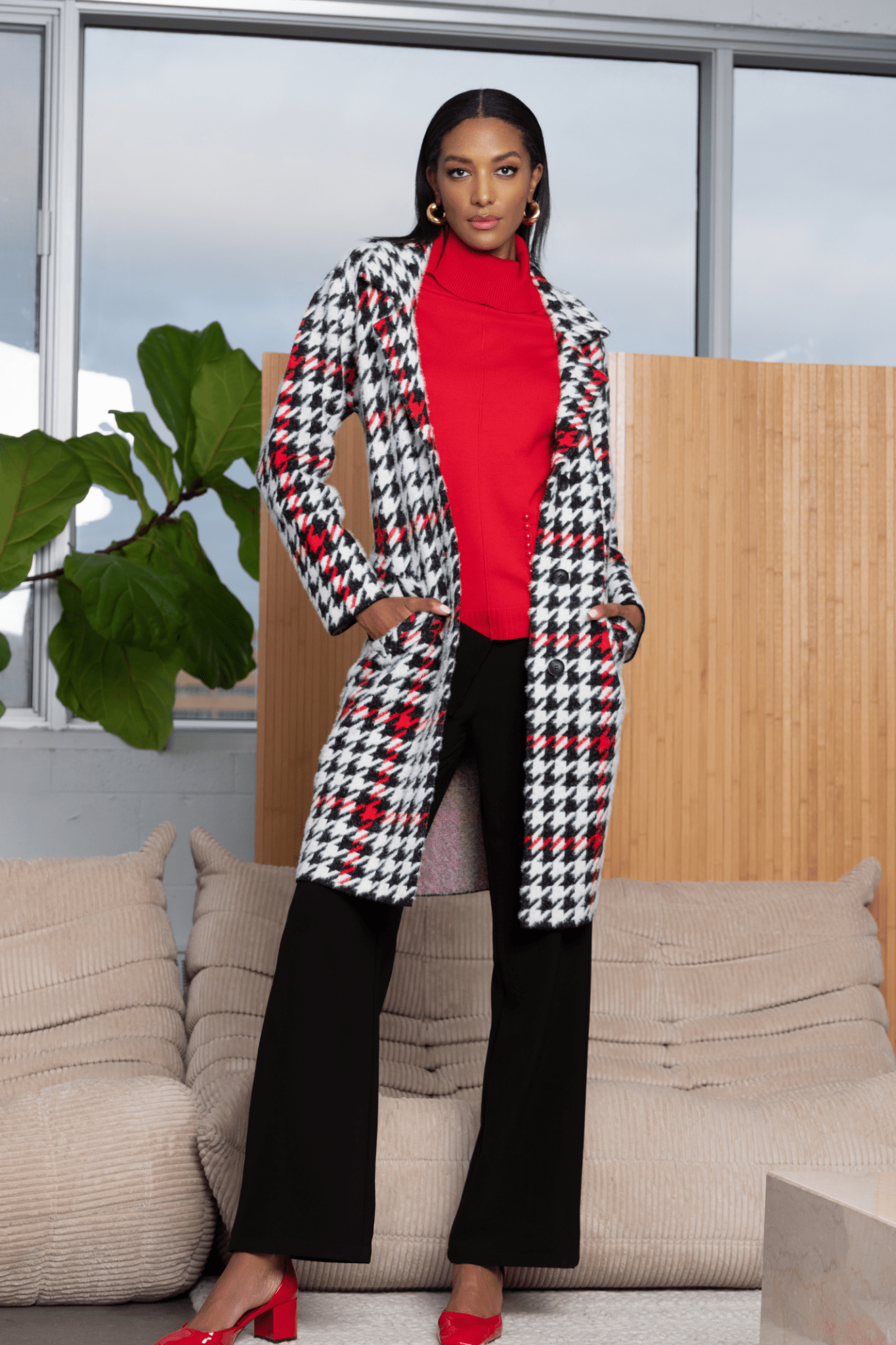 Woman wearing a houndstooth coat over a red sweater, standing in a modern living room.