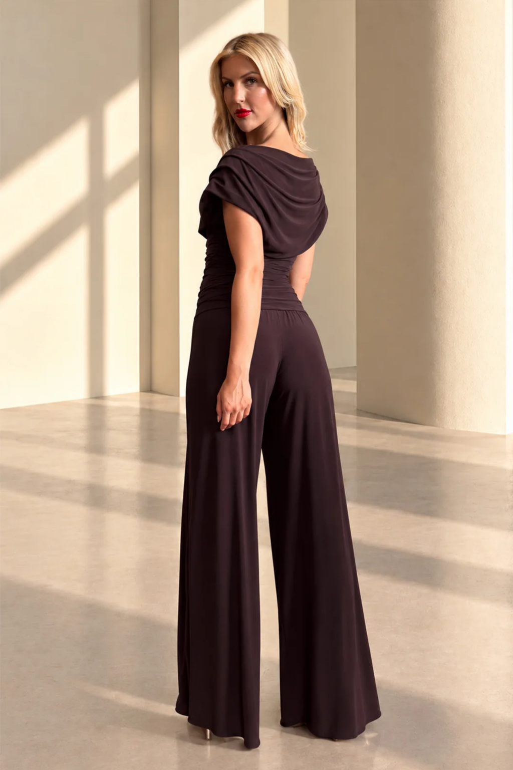 Woman wearing a dark brown draped jumpsuit standing in a sunlit room