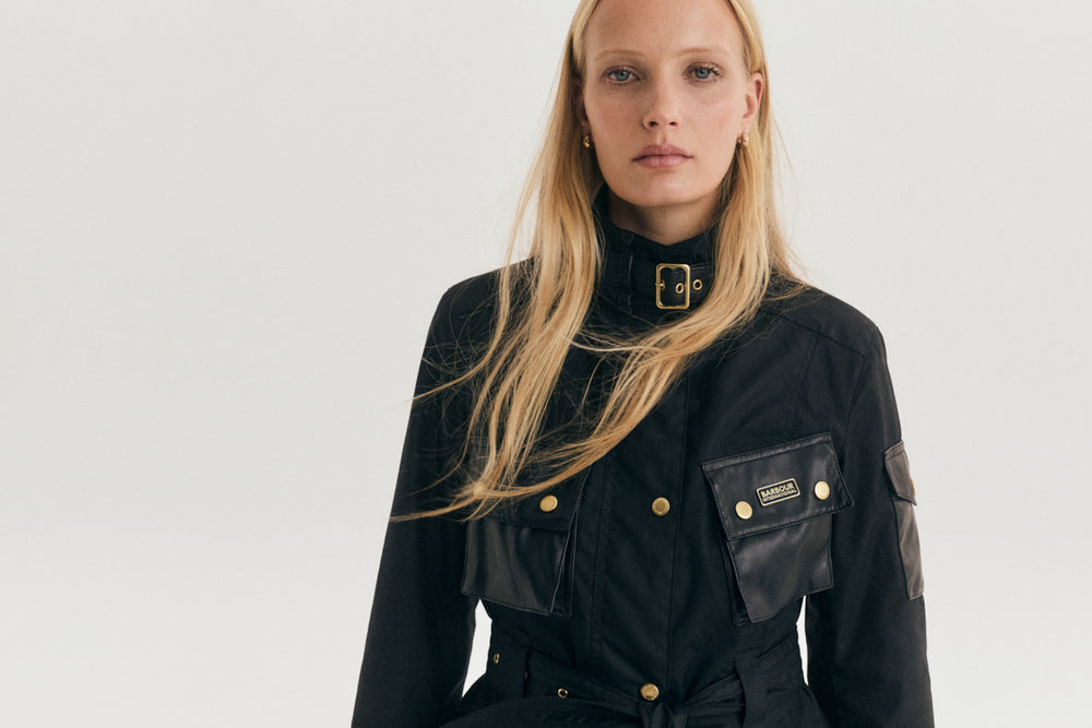 A blonde model wearing a black Barbour jacket with gold buttons and a high collar, photographed against a light gray studio background.
