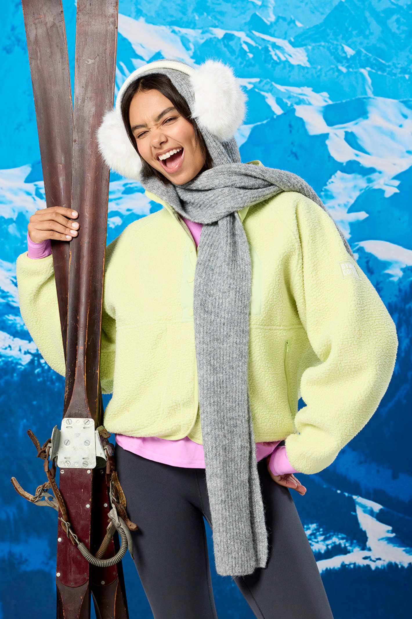 Woman wearing Gym + Coffee Horizon Layer Up Jacket in in Lime Sorbet by Gym+Coffee holding skis, shop Gym+Coffee at Dotique