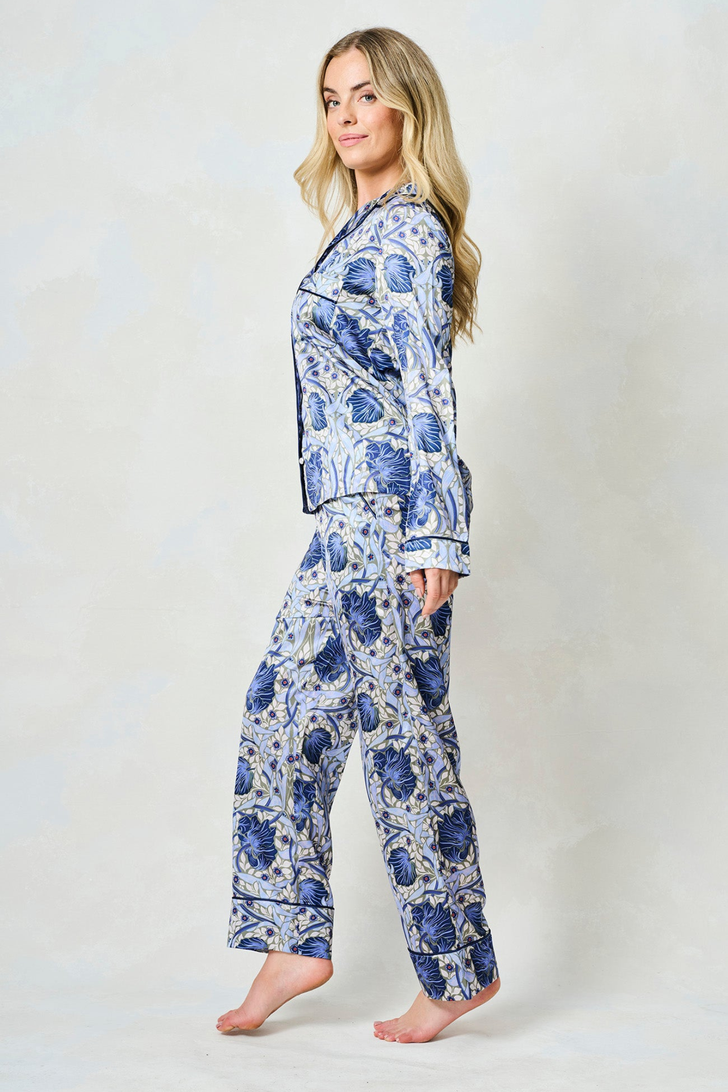 Woman standing in blue and white floral patterned satin pyjama set with long sleeves and straight-leg trousers against light background