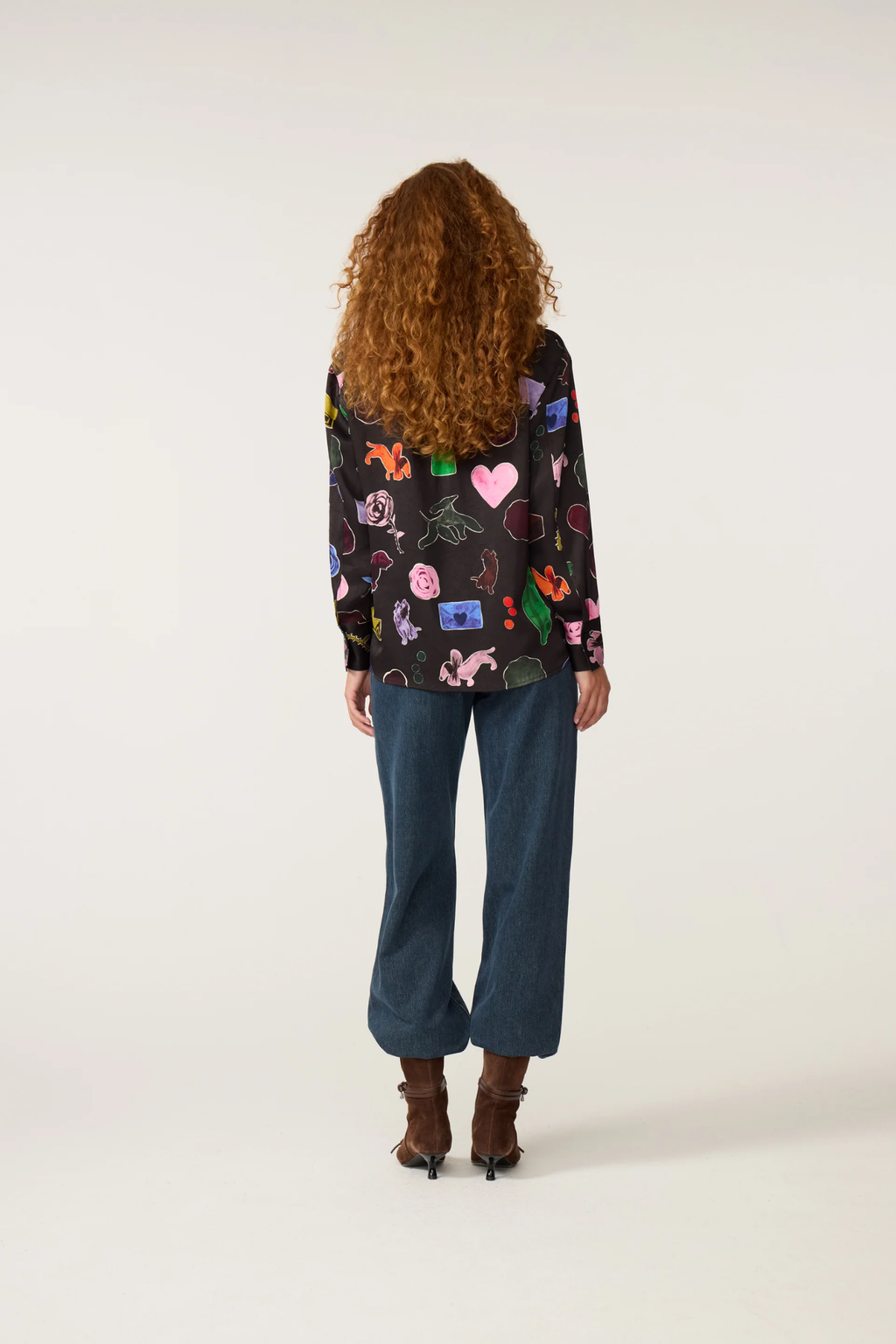 Person seen from behind wearing a colorful embroidered jacket, blue jeans, and brown ankle boots against a white background