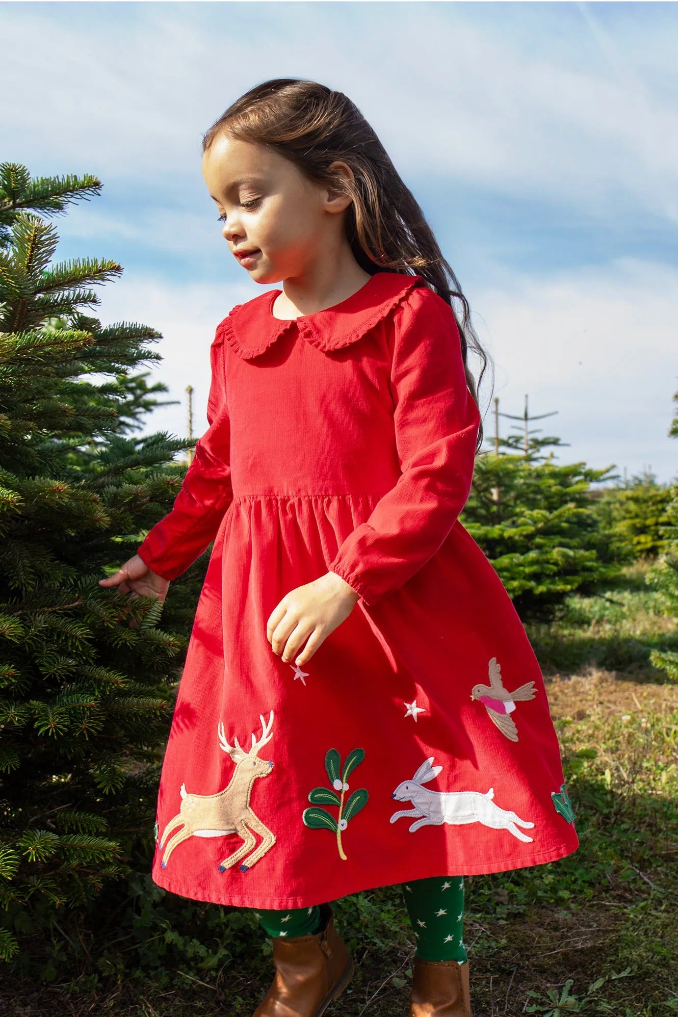 Young girl in a red dress with animal appliqués standing next to a Christmas tree.