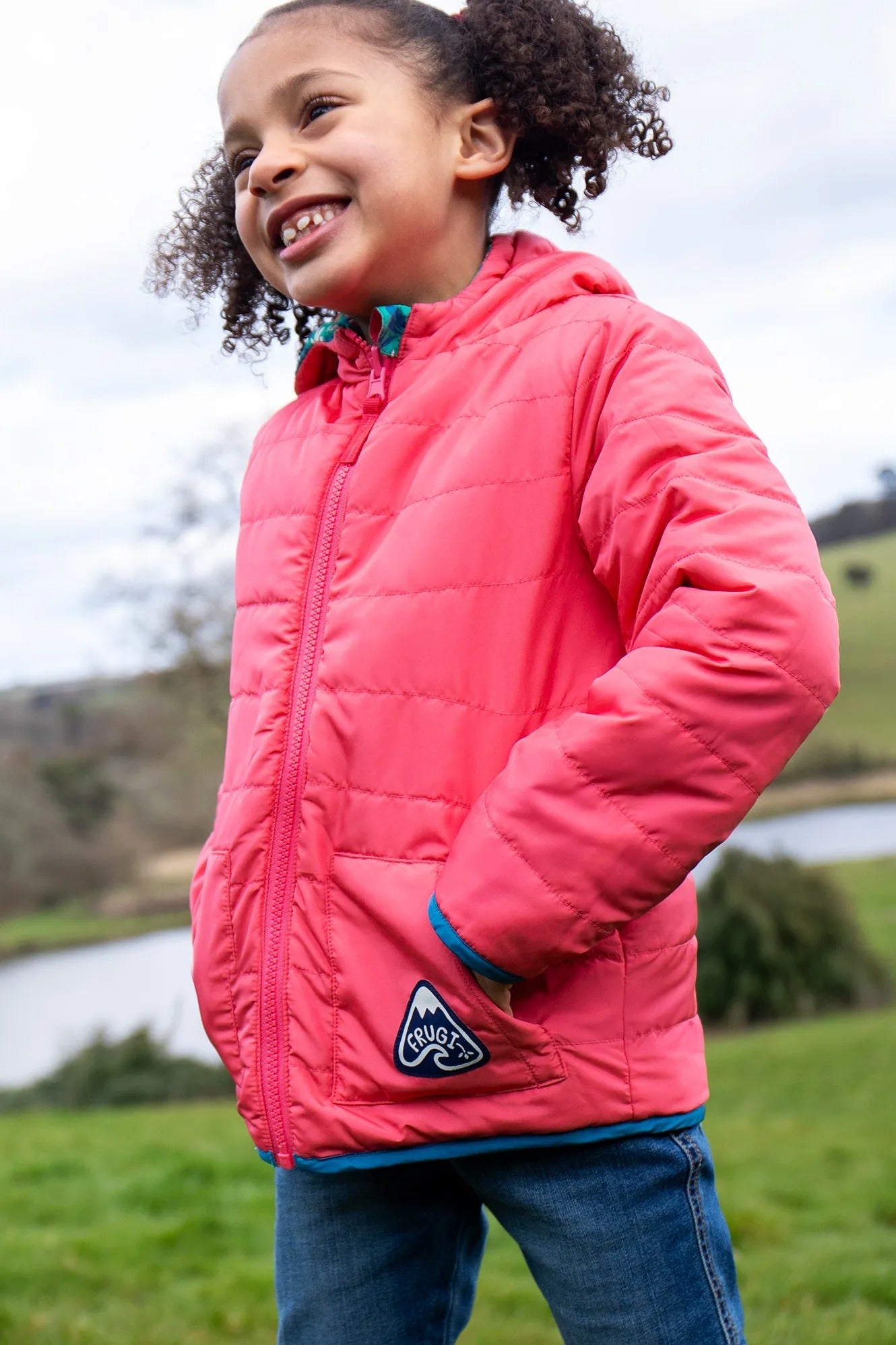 Child wearing a pink jacket with a logo, standing outdoors near a body of water.