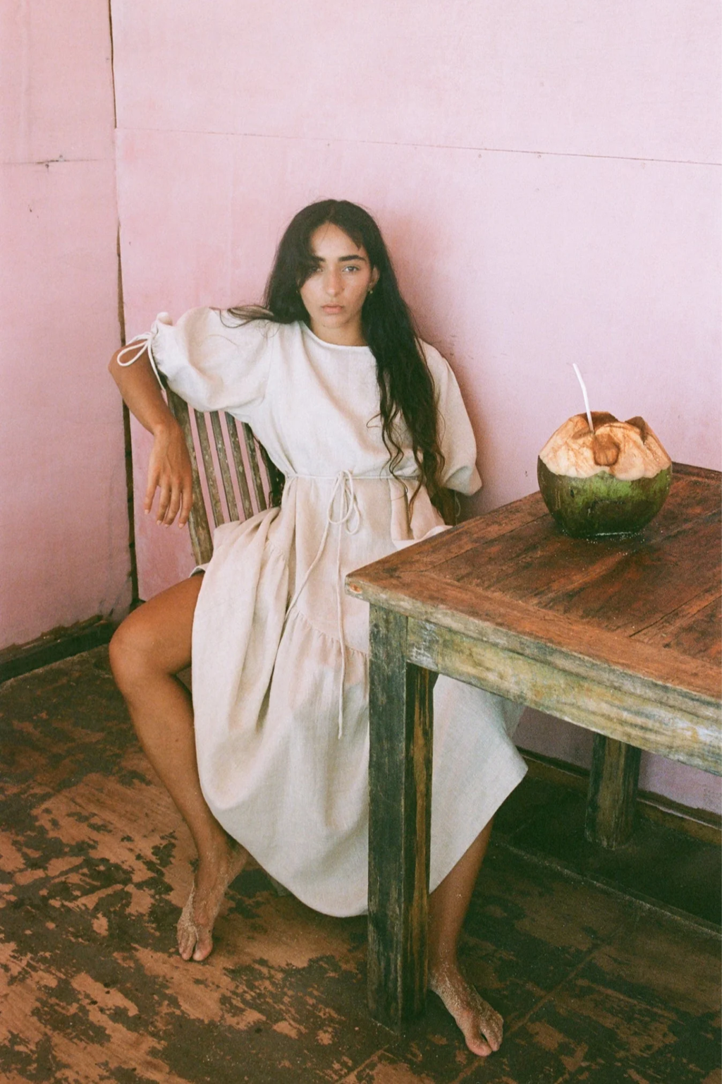 Lilly Pilly LPSS22-D002 Charlotte Linen Dress Oatmeal by Lilly Pilly modeled barefoot beside wooden table Shop Lilly Pilly at Dotique