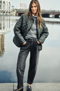 A woman stands by a waterfront wearing a dark quilted jacket over a gray sweater and dark jeans, with a bridge and water in the background. Shop MAC Jeans at Dotique