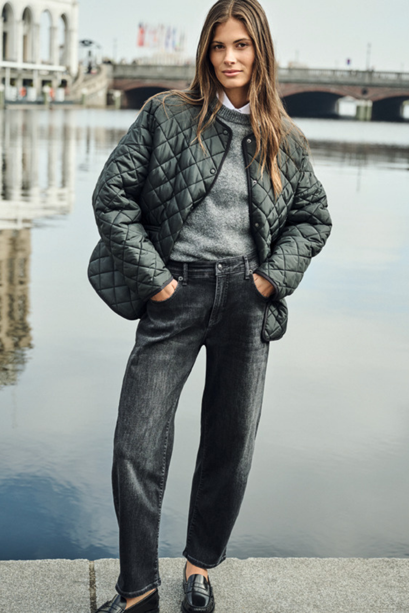 A woman stands by a waterfront wearing a dark quilted jacket over a gray sweater and dark jeans, with a bridge and water in the background. Shop MAC Jeans at Dotique