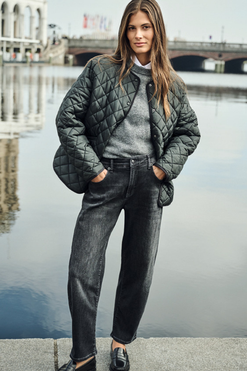 A woman stands by a waterfront wearing a dark quilted jacket over a gray sweater and dark jeans, with a bridge and water in the background. Shop MAC Jeans at Dotique