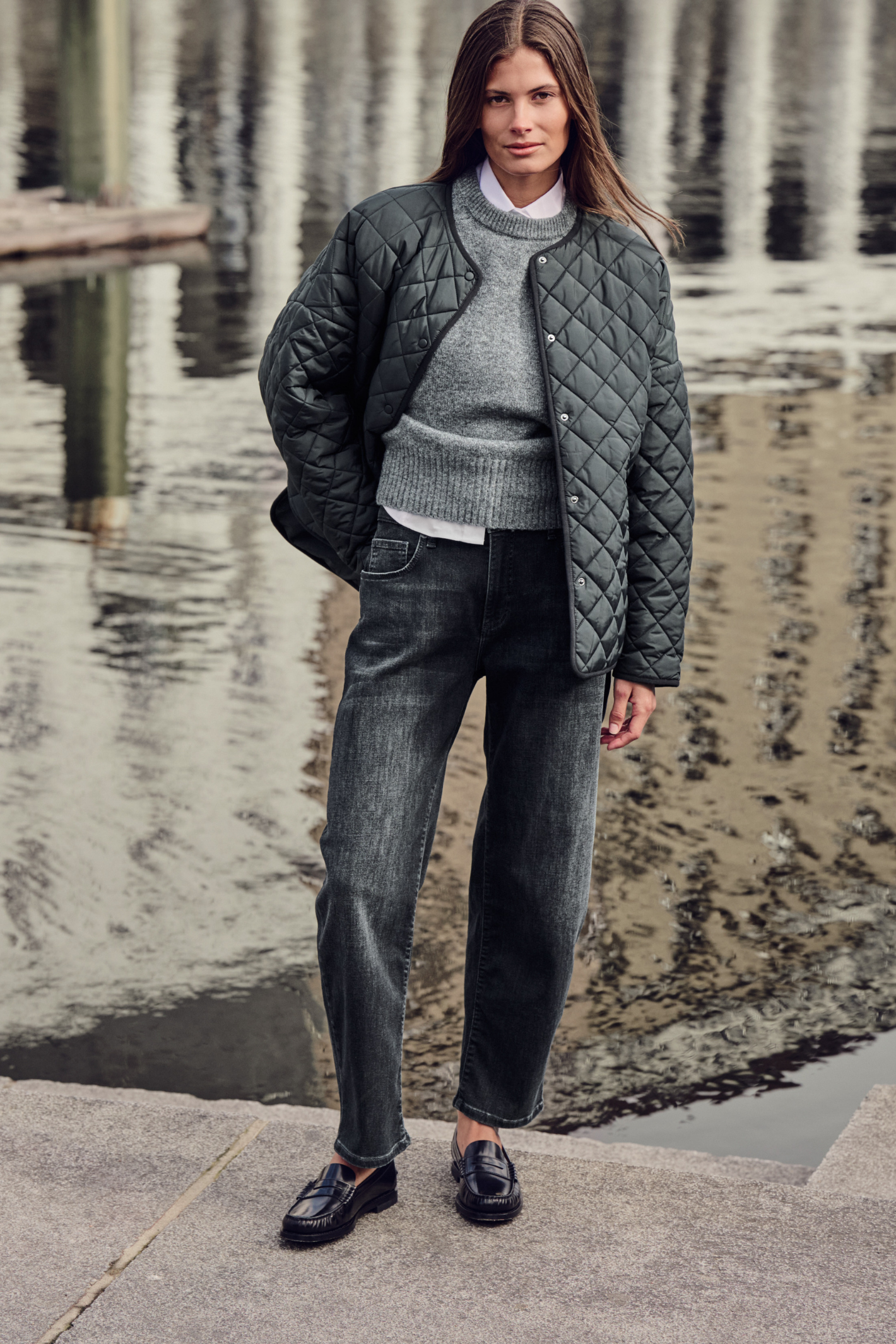 A woman stands outdoors by a reflective body of water wearing a dark quilted jacket over a gray knit sweater, dark jeans, and black loafers. Shop MAC Jeans at Dotique