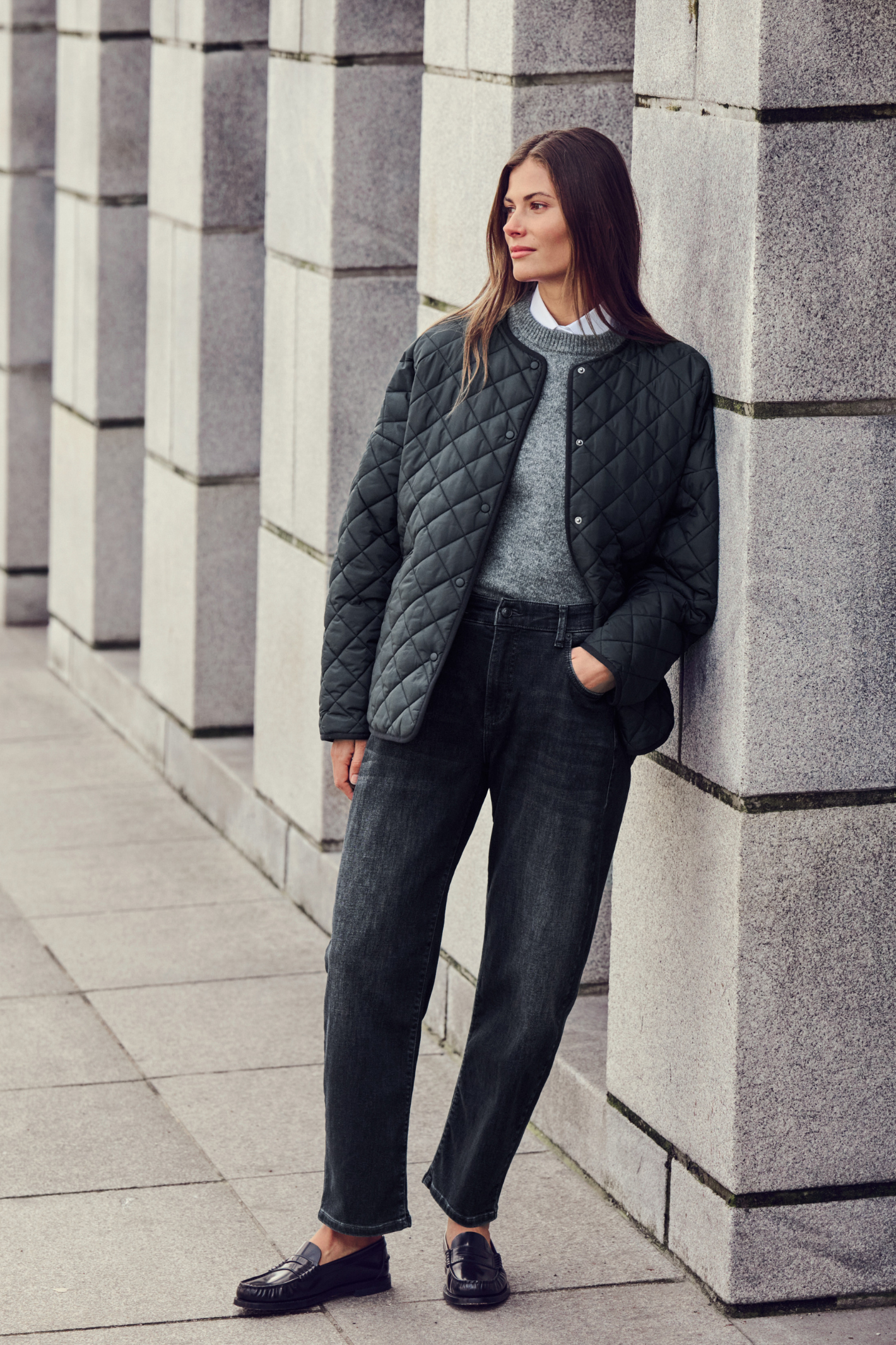 A woman stands against a gray stone column wall wearing a black quilted jacket over a gray sweater, dark jeans, and black loafers.Shop MAC Jeans at Dotique