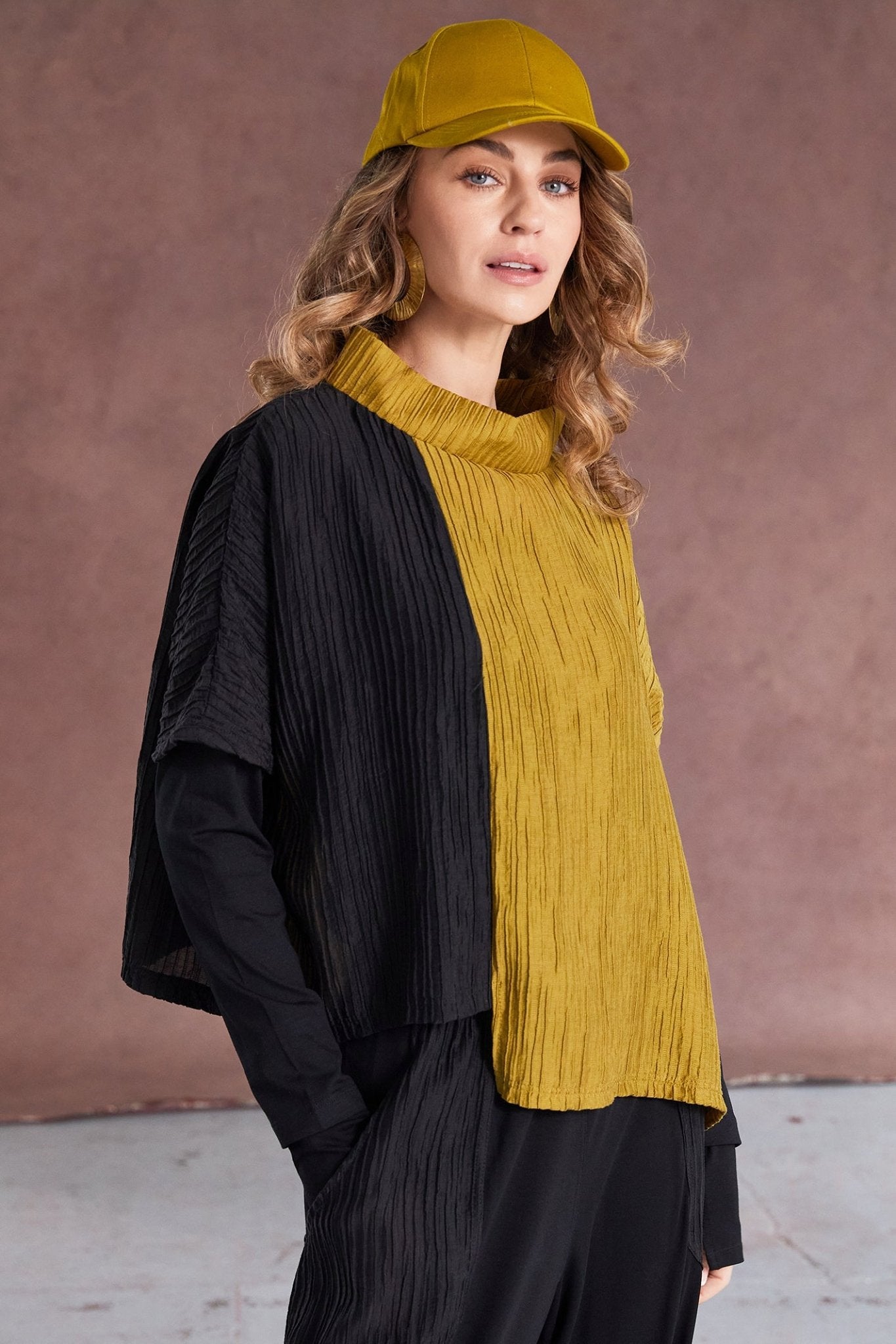 Woman wearing a mustard yellow top and cap with a black jacket against a brown background