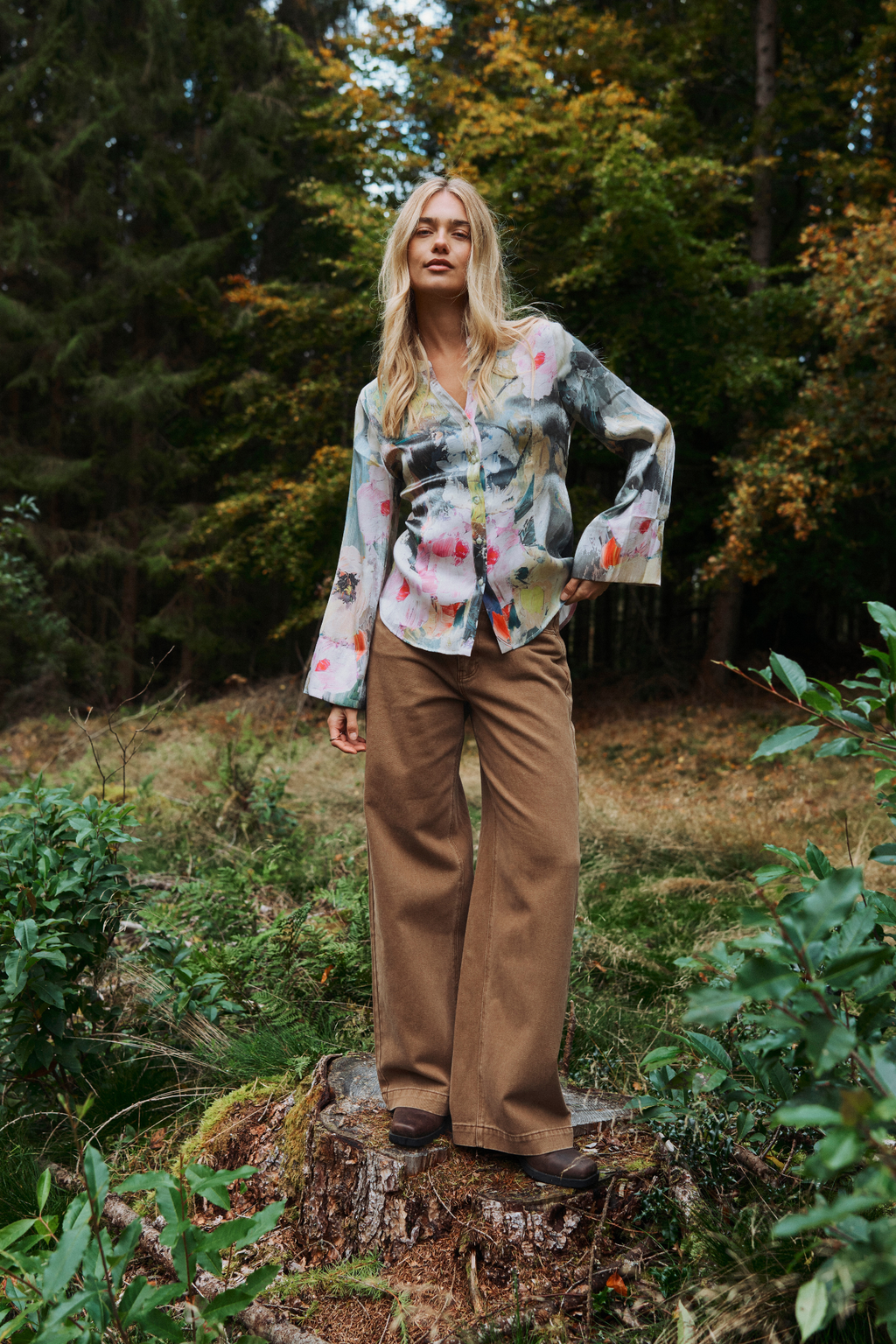 A woman stands on a tree stump in a forest setting, wearing a long-sleeve button-up shirt with a multicolor floral print and brown wide-leg pants.