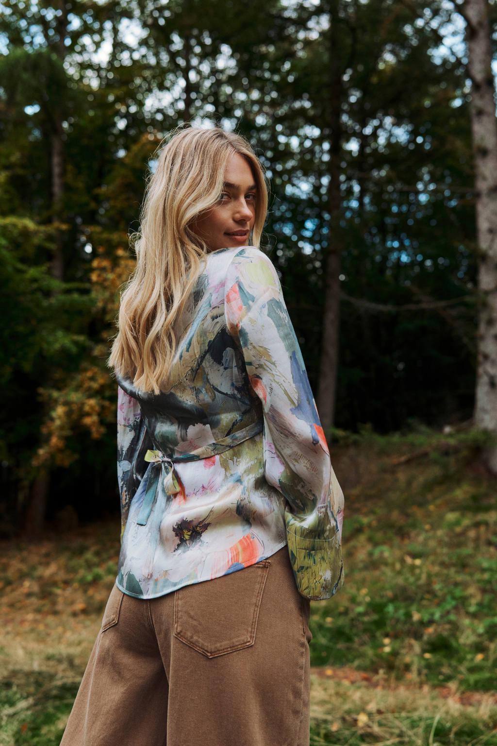 A woman standing outdoors in a wooded setting, wearing a long-sleeve tie-front wrap top with a multicolor abstract floral print and brown wide-leg pants.