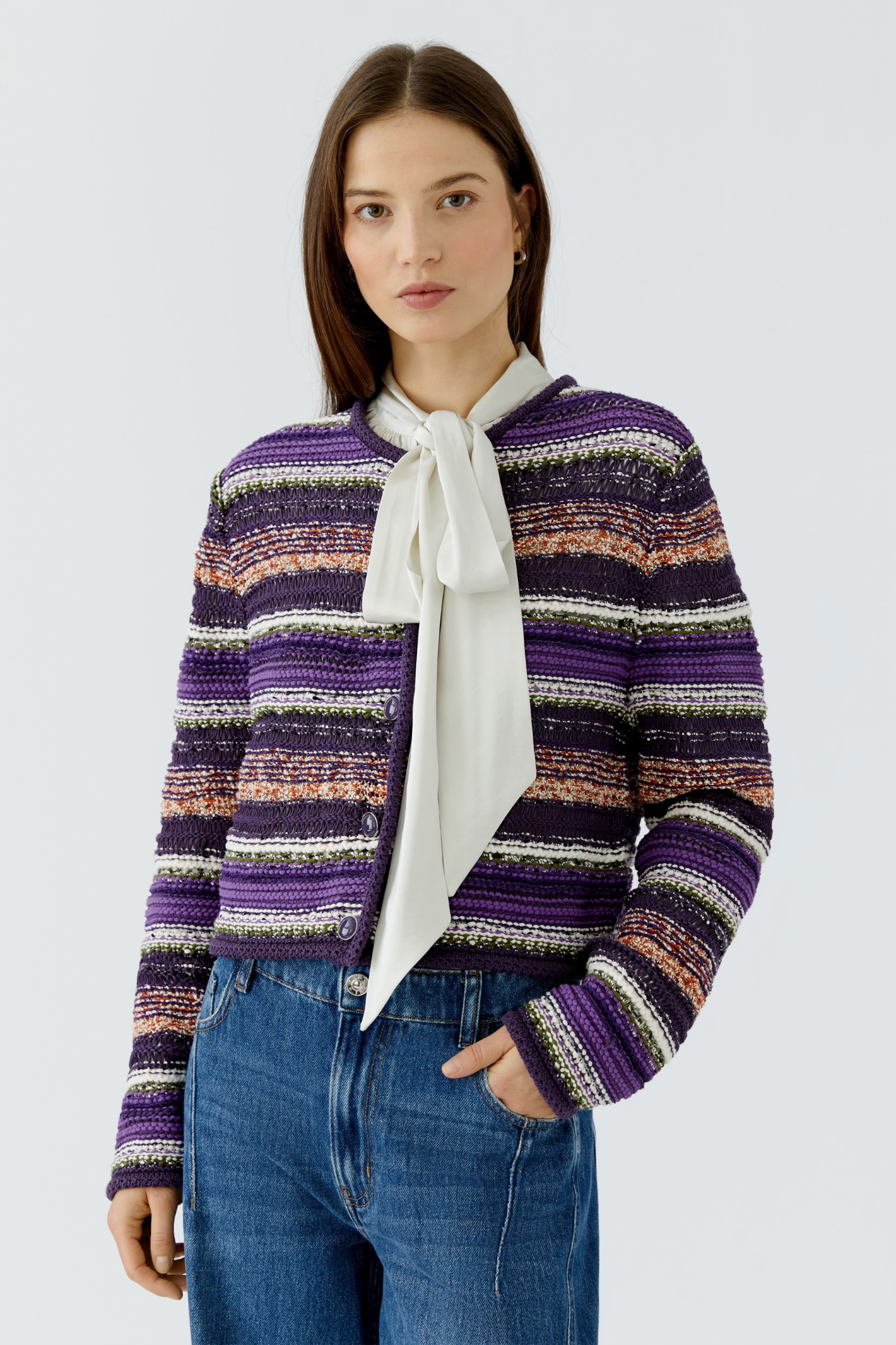 A woman modeling a cropped, textured knit cardigan with horizontal stripes in purple, white, and orange-brown, worn over a white bow-neck blouse and blue jeans against a light gray studio background.