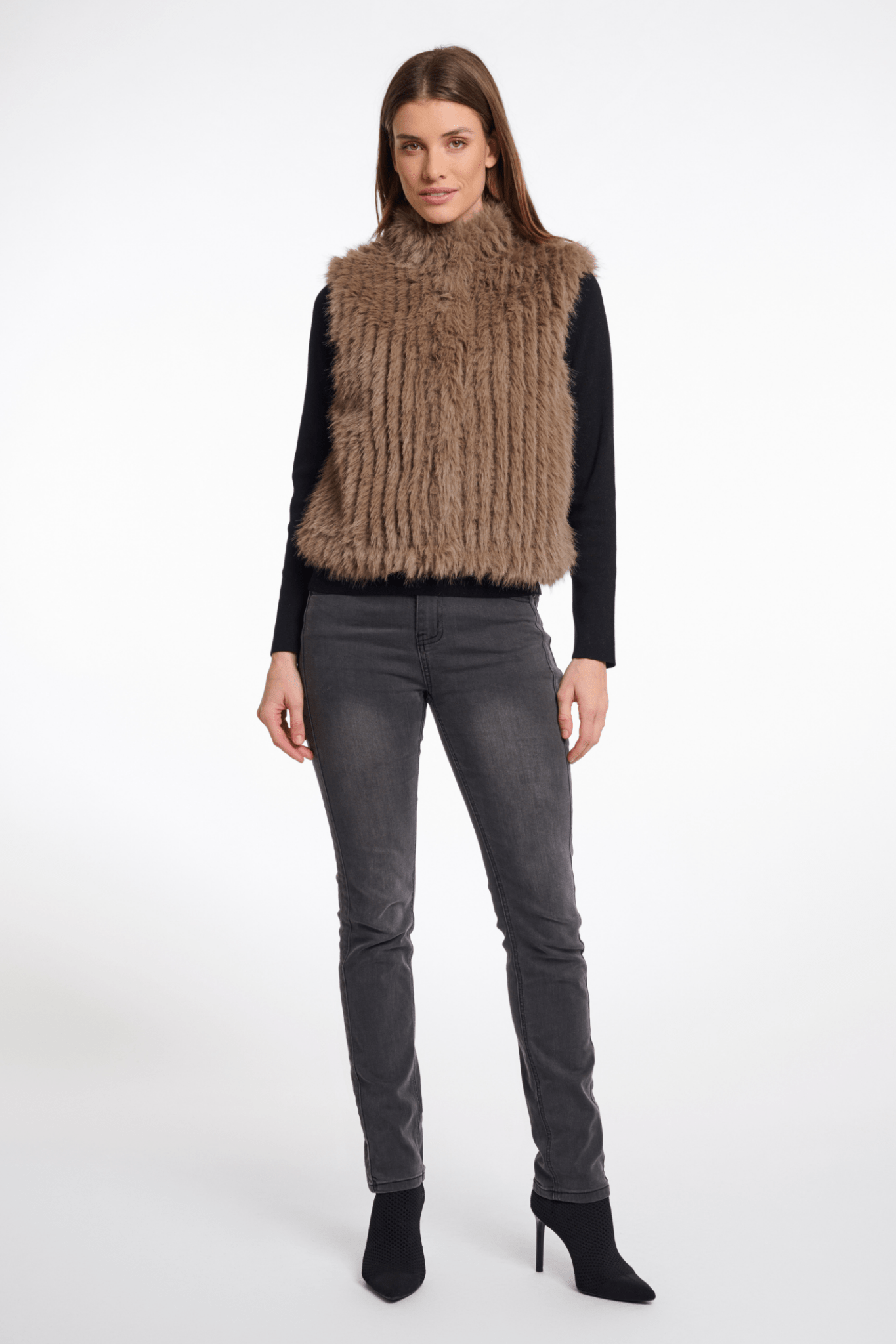 Woman wearing a brown fur vest over a black long-sleeve shirt with gray jeans on a white background